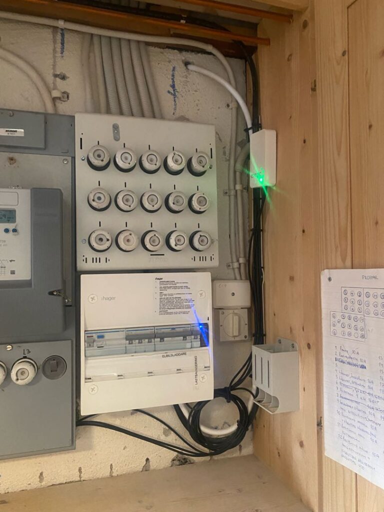 Bild som visar en elektrisk brytarpanel monterad på en vägg, med flera säkringar och ledningar synliga. I anslutning till panelen finns en digital enhet med ett lysande grönt ljus. Ett papper med handskrivna anteckningar klistras på en träyta bredvid panelen i Falun, där stolpe laddbox-installationer är vanliga.