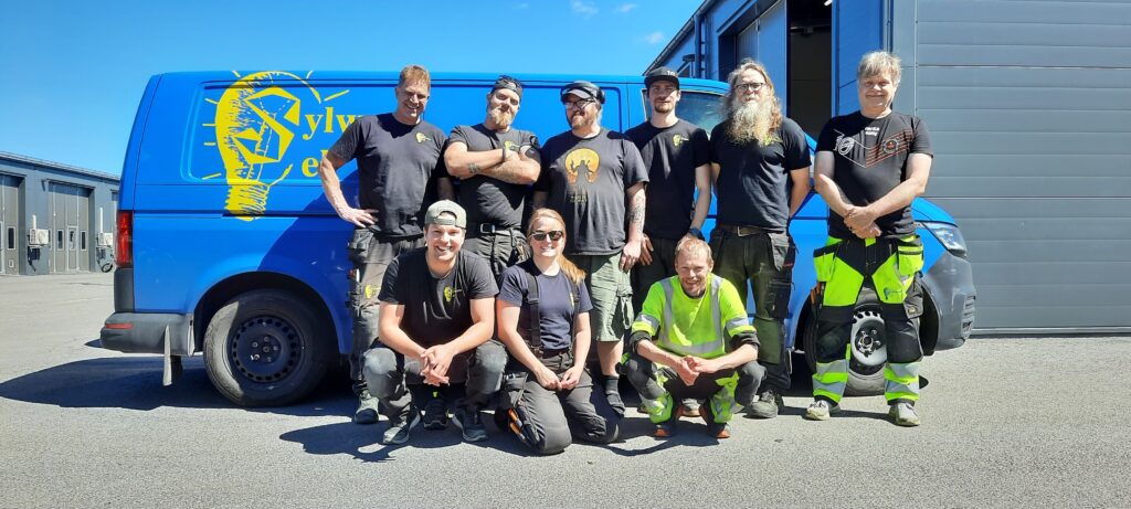 Personal I Sylwans El poserar framför en blå bil av märket Volkswagen transporter med en gul sylwans el logga till vänster i bild.